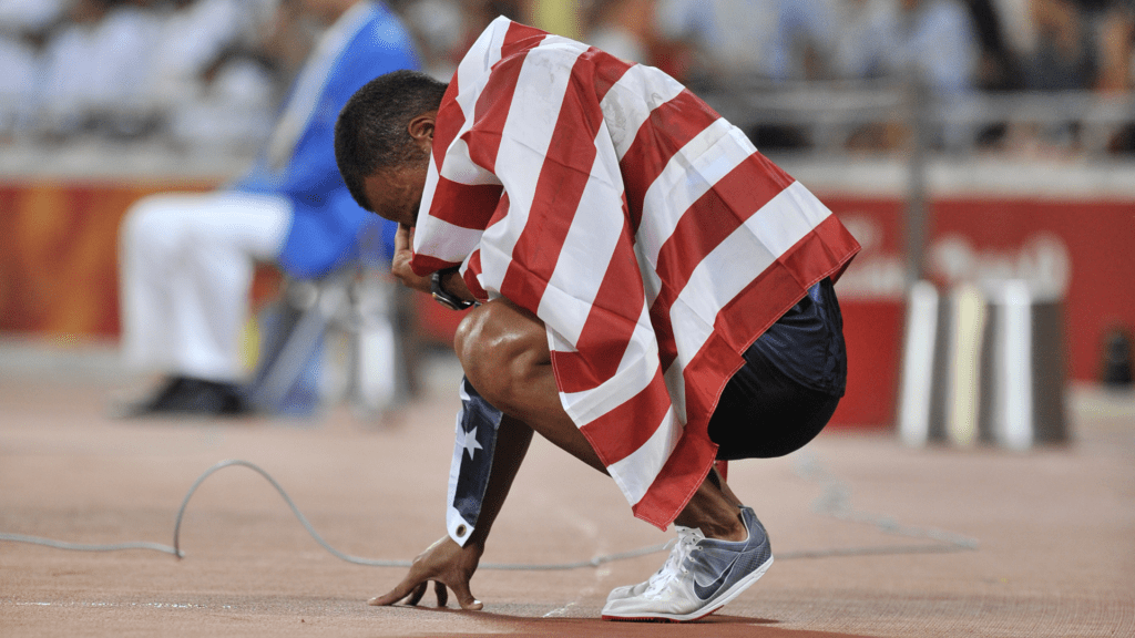 Bryan Clay kneeling on the track wrapped in an American flag after winning the 2008 Beijing Olympic decathlon